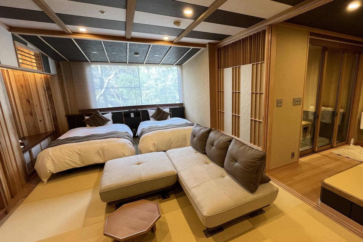 A hotel room with a sectional sofa, two single beds, a small table, and a sliding glass door leading to a bathroom.