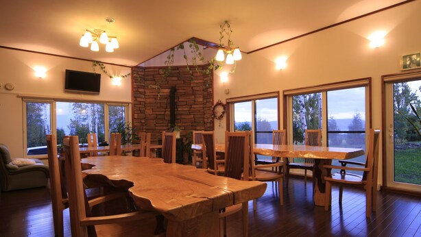 A spacious dining area with a large wooden table and chairs, surrounded by large windows offering views of the outdoors. - Pension ASHITAYA (Furano)