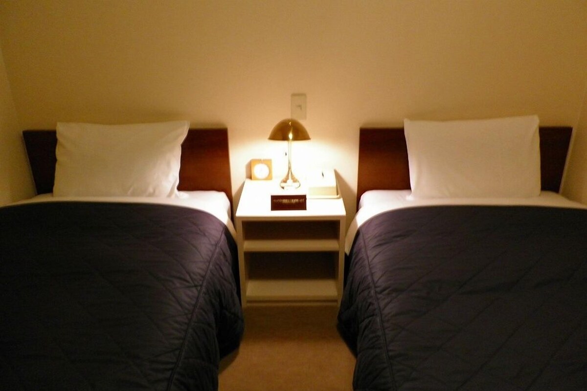 A hotel room with two beds, each with a headboard, a bedside table with a lamp, and a small card on the table.