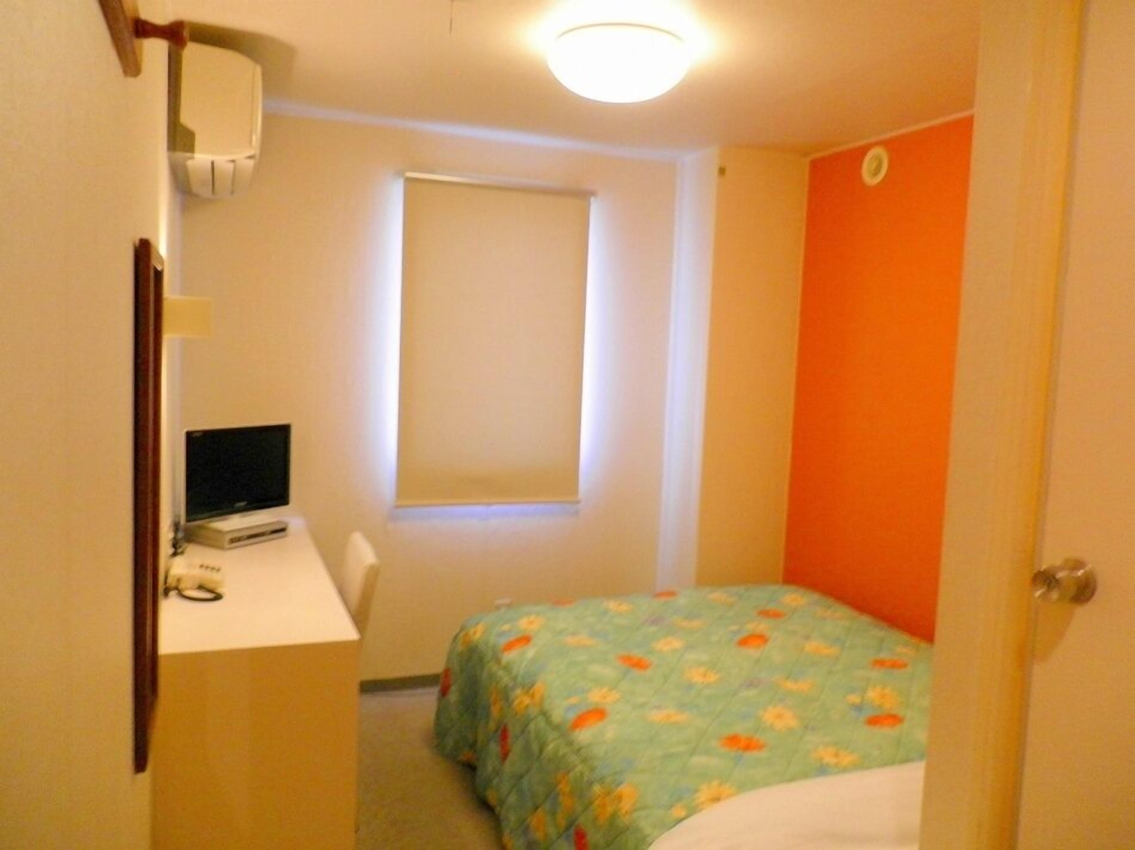 A small room with a bed, a desk with a computer, a window, and an orange wall.