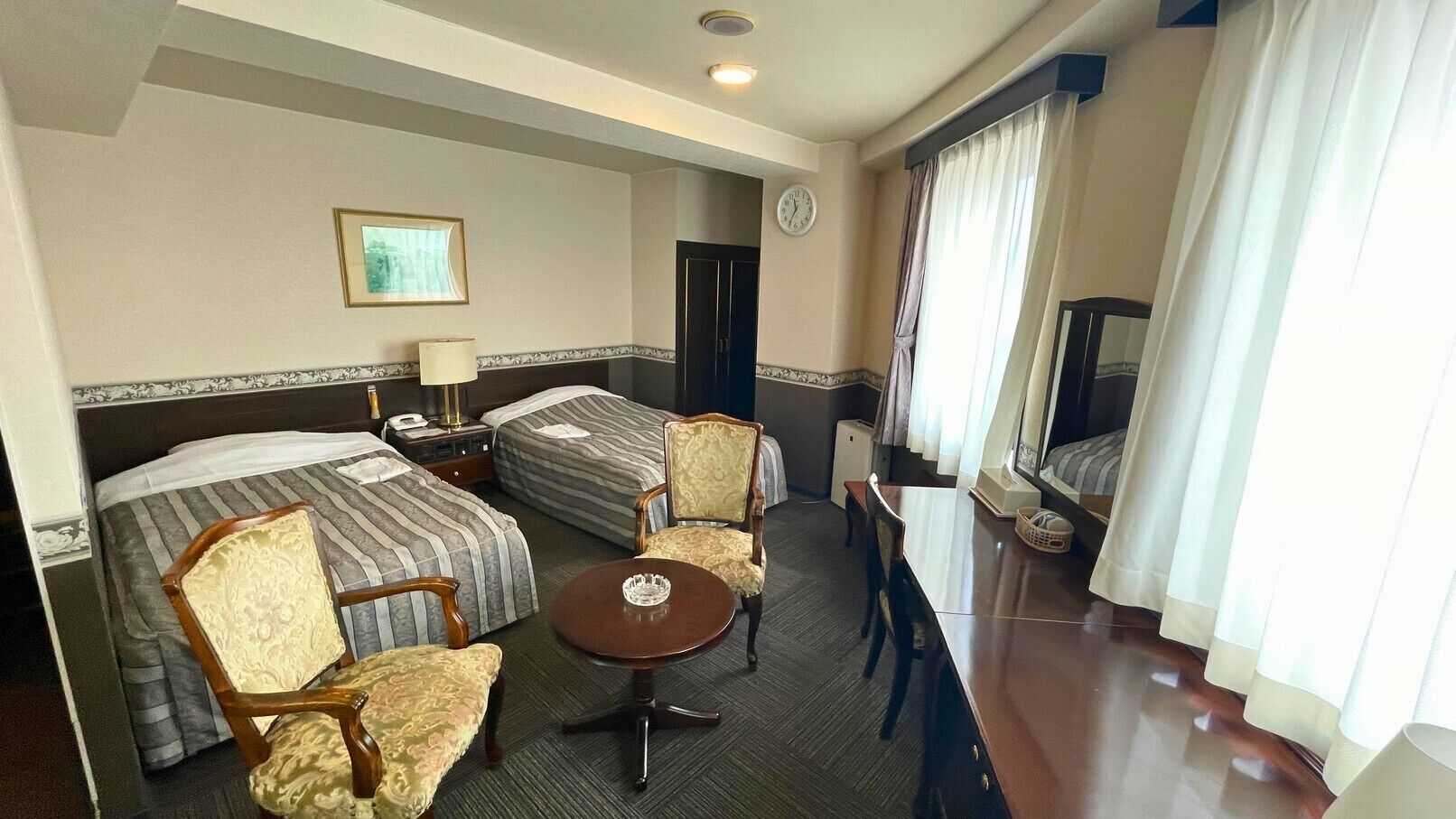 A hotel room with two beds, a small table, a chair, a desk, a television, and a clock on the wall.