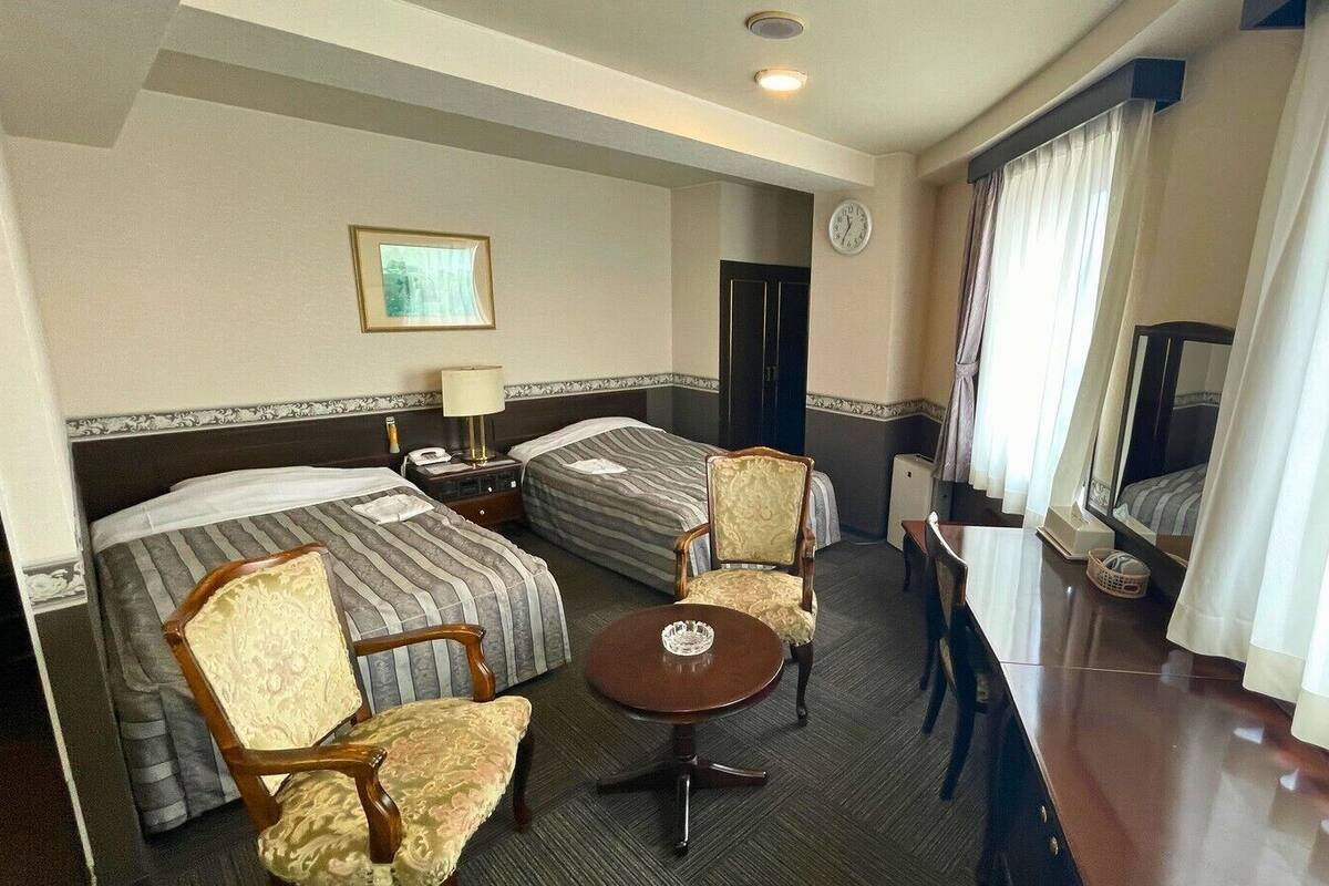 A hotel room with two beds, a small table, a chair, a desk, a television, and a clock on the wall.