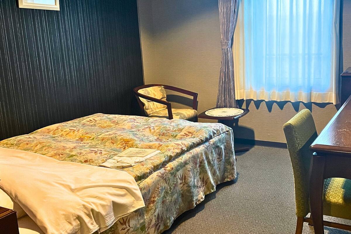 A hotel room with a bed, a chair, a small table, and a window with curtains.