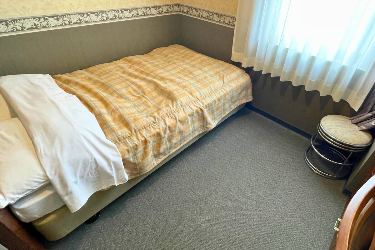 A small, single-bed room with a grey carpet, a plaid bedspread, and a small round stool.