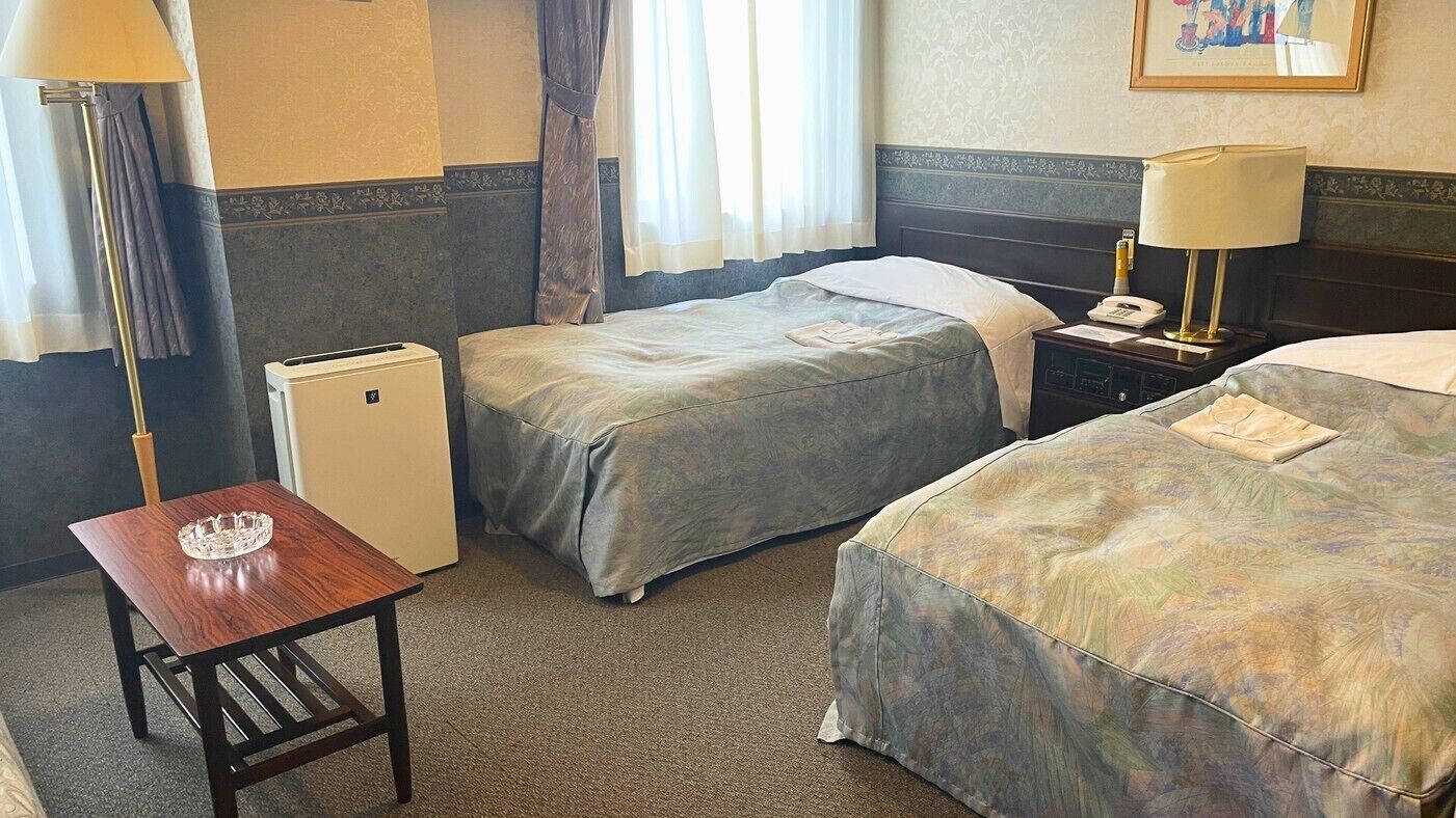 A hotel room with two beds, a desk, a lamp, a nightstand, and a small air conditioning unit.