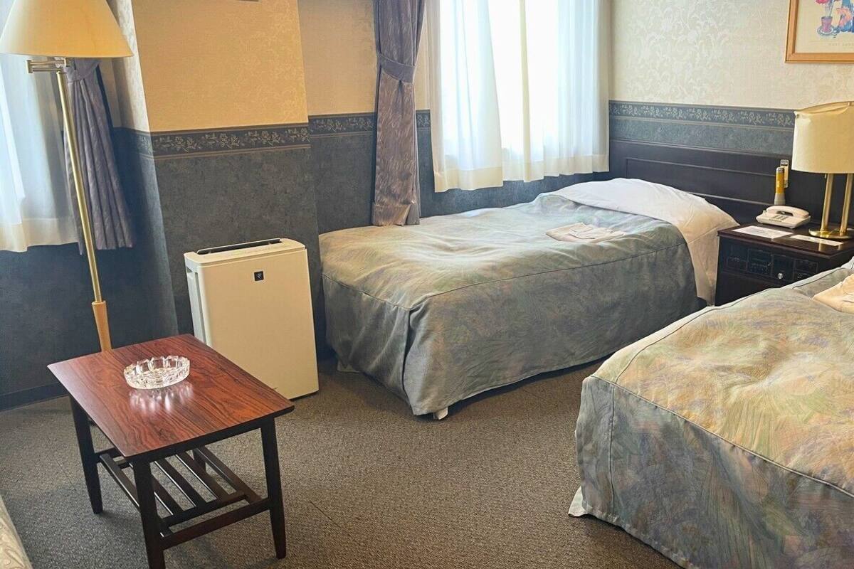 A hotel room with two beds, a desk, a lamp, a nightstand, and a small air conditioning unit.