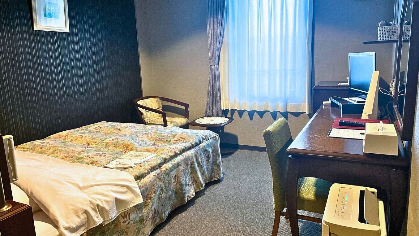 A hotel room with a bed, a desk with a computer, a chair, and a window with curtains.