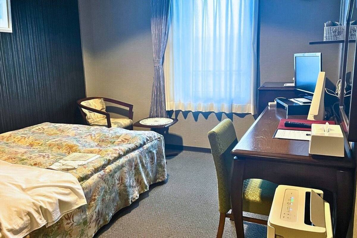 A hotel room with a bed, a desk with a computer, a chair, and a window with curtains.