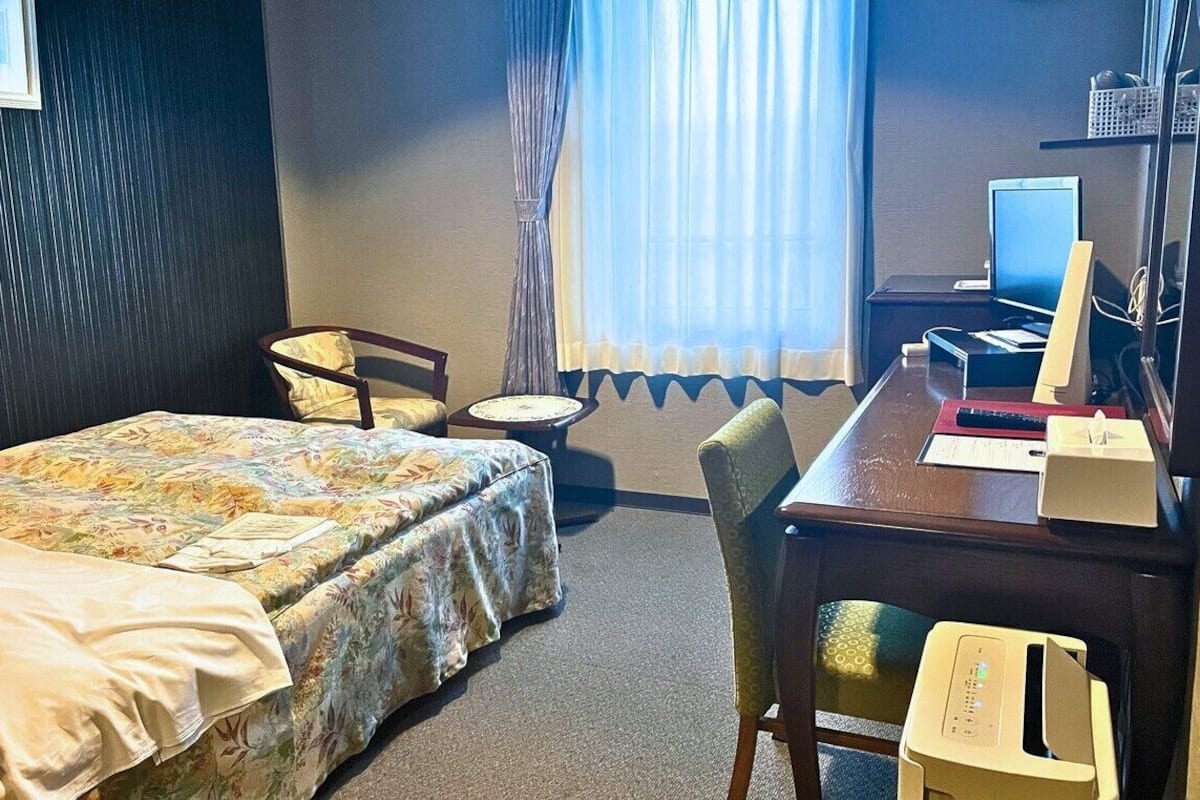 A hotel room with a bed, a desk with a computer, a chair, and a window with curtains.