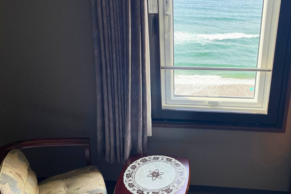 A room with a view of the ocean through a window, a chair, and a table with a doily.