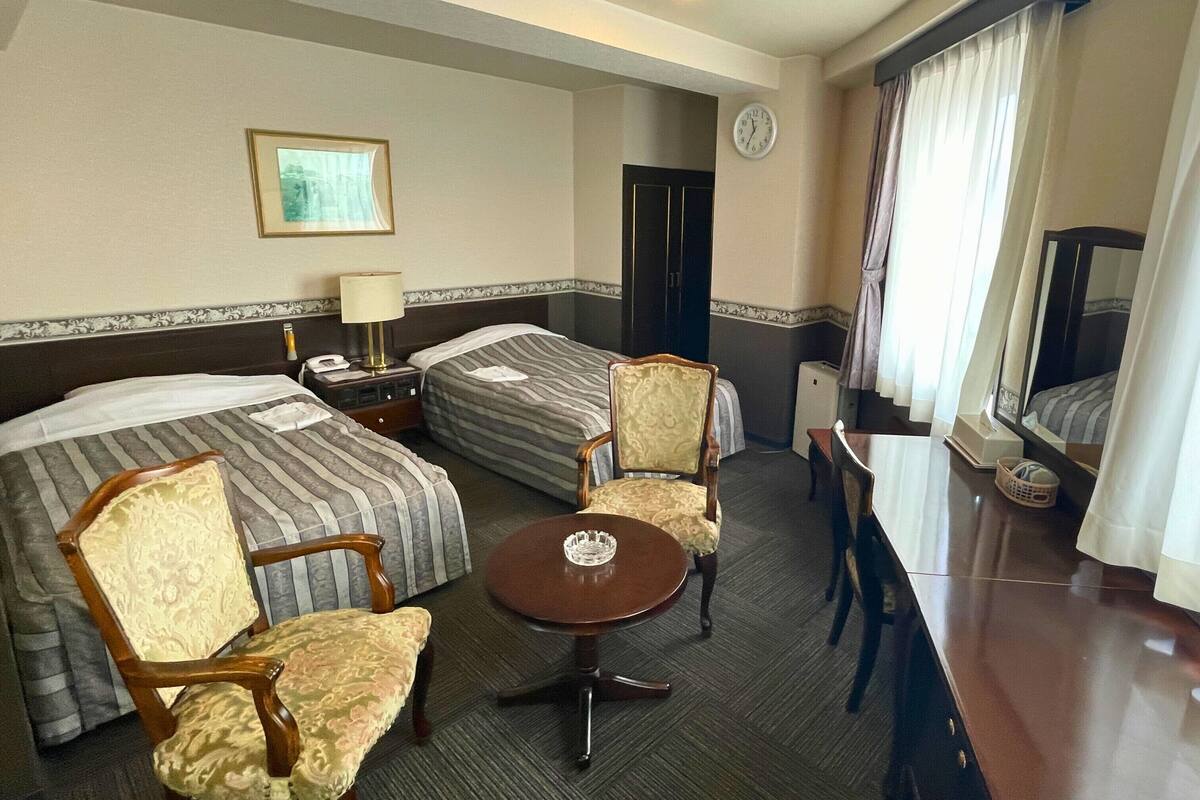 A hotel room with two beds, a desk, a chair, a small table, a clock, and a window with curtains.
