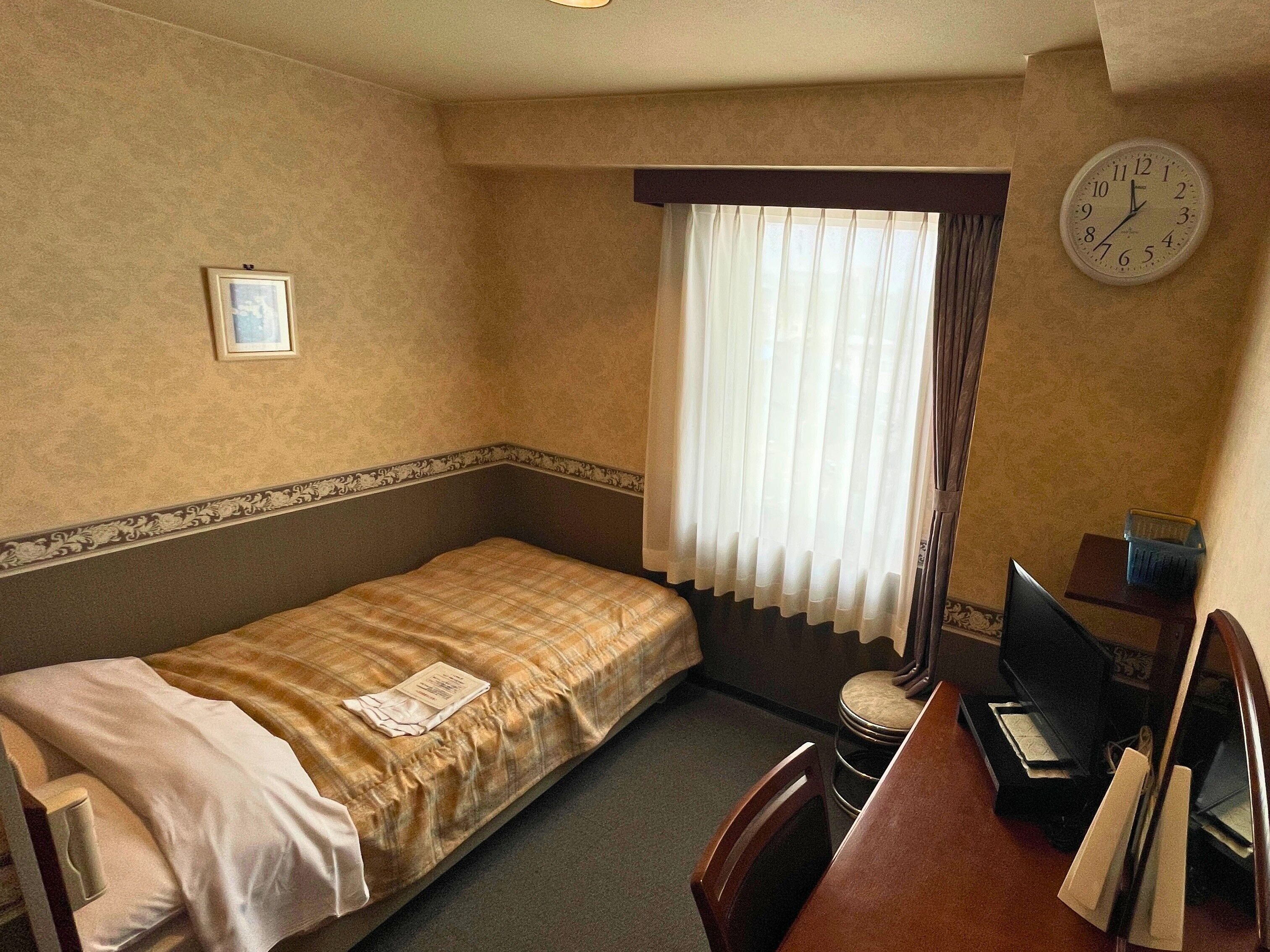 A small, single-bed room with a desk, chair, and a clock on the wall.