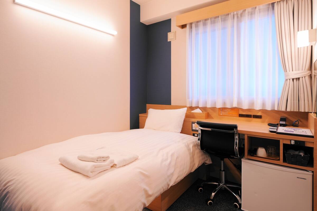 Ein Hotelzimmer mit einem Bett, einem Schreibtisch mit Stuhl, einer kleinen Kühlbox und einem Fenster mit Vorhängen.