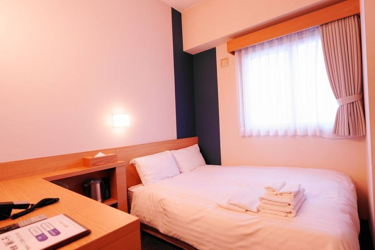 Ein Hotelzimmer mit Bett, Schreibtisch, Lampe und einem fensterbelaubten Fenster.