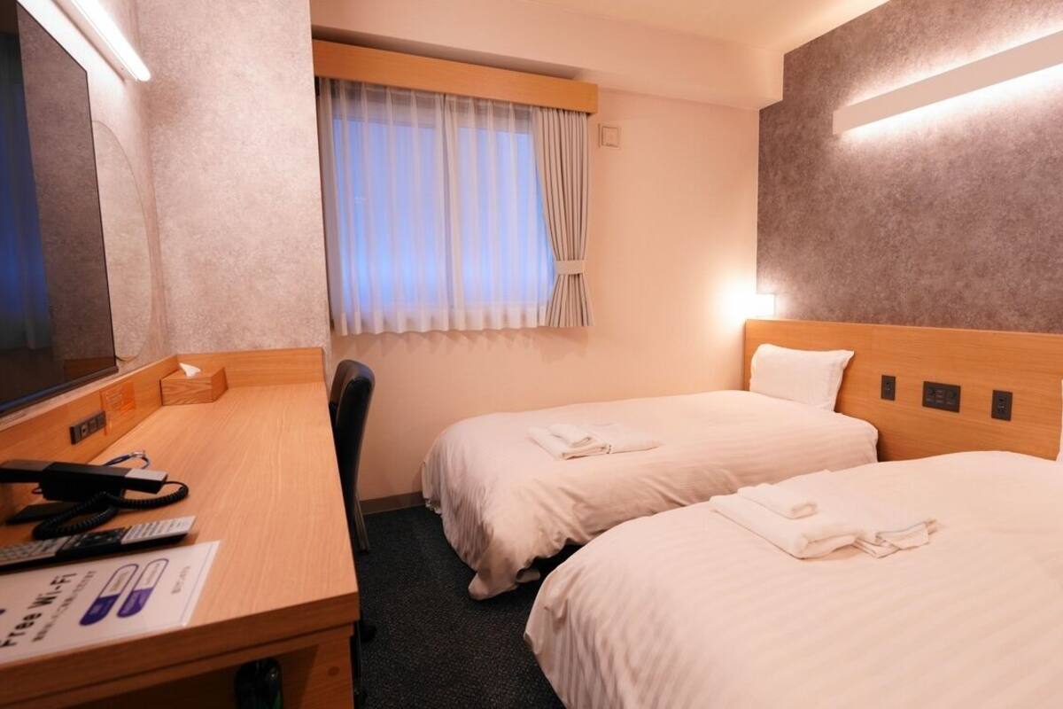 Ein Hotelzimmer mit zwei Betten, einem Schreibtisch mit Telefon und einem fensterbepflanzten Fenster.