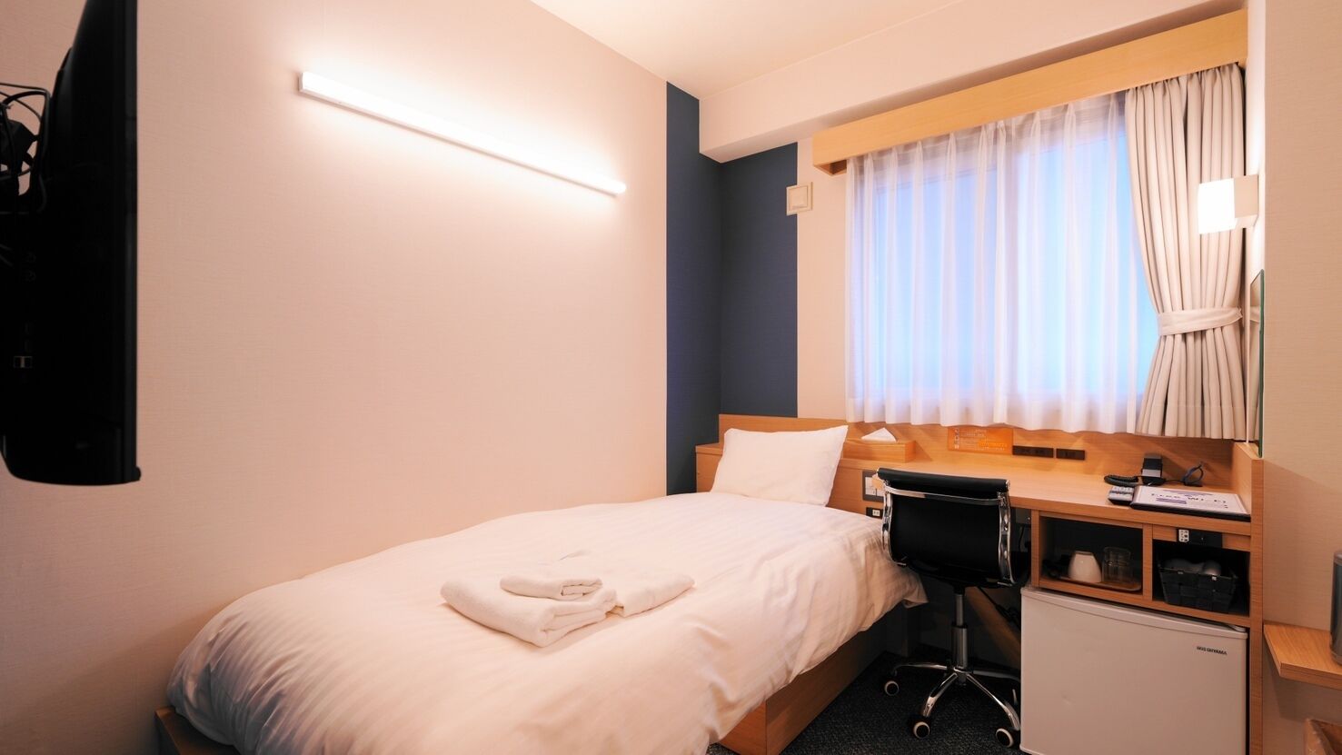 A hotel room with a bed, a desk, a chair, a small refrigerator, a flat-screen TV, and a window with curtains.