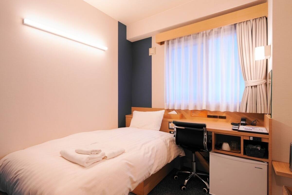 Ein Hotelzimmer mit Bett, Schreibtisch, Stuhl, kleinem Kühlschrank, Flachbildfernseher und einem Fenster mit Vorhängen.