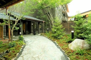 A stone pathway leading to a traditional building with a tiled roof, surrounded by lush greenery and rocks. - Konoha (Kusatsu)