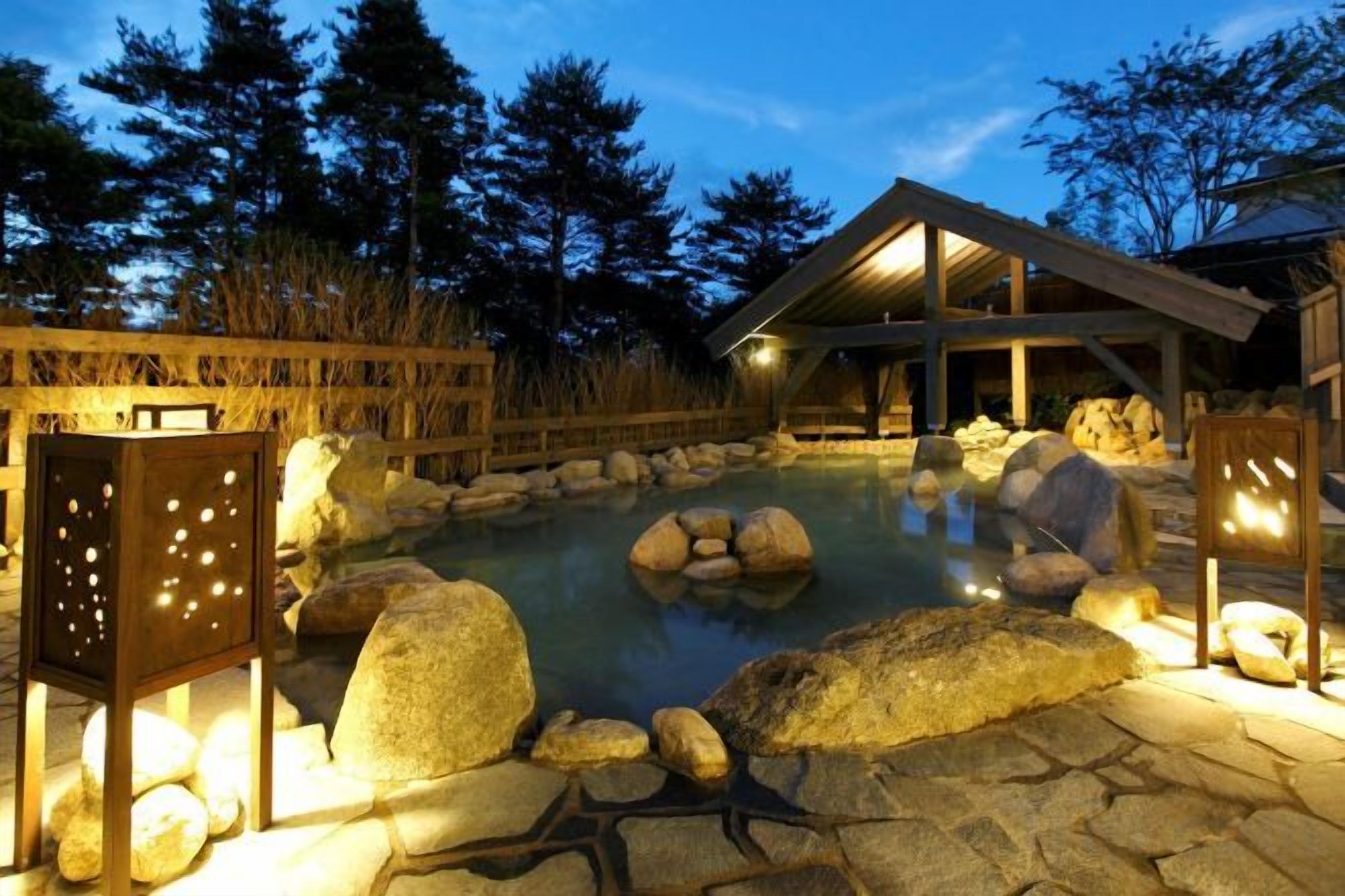 A heated outdoor pool with rocks and a wooden deck, surrounded by trees and a building with a sloped roof.