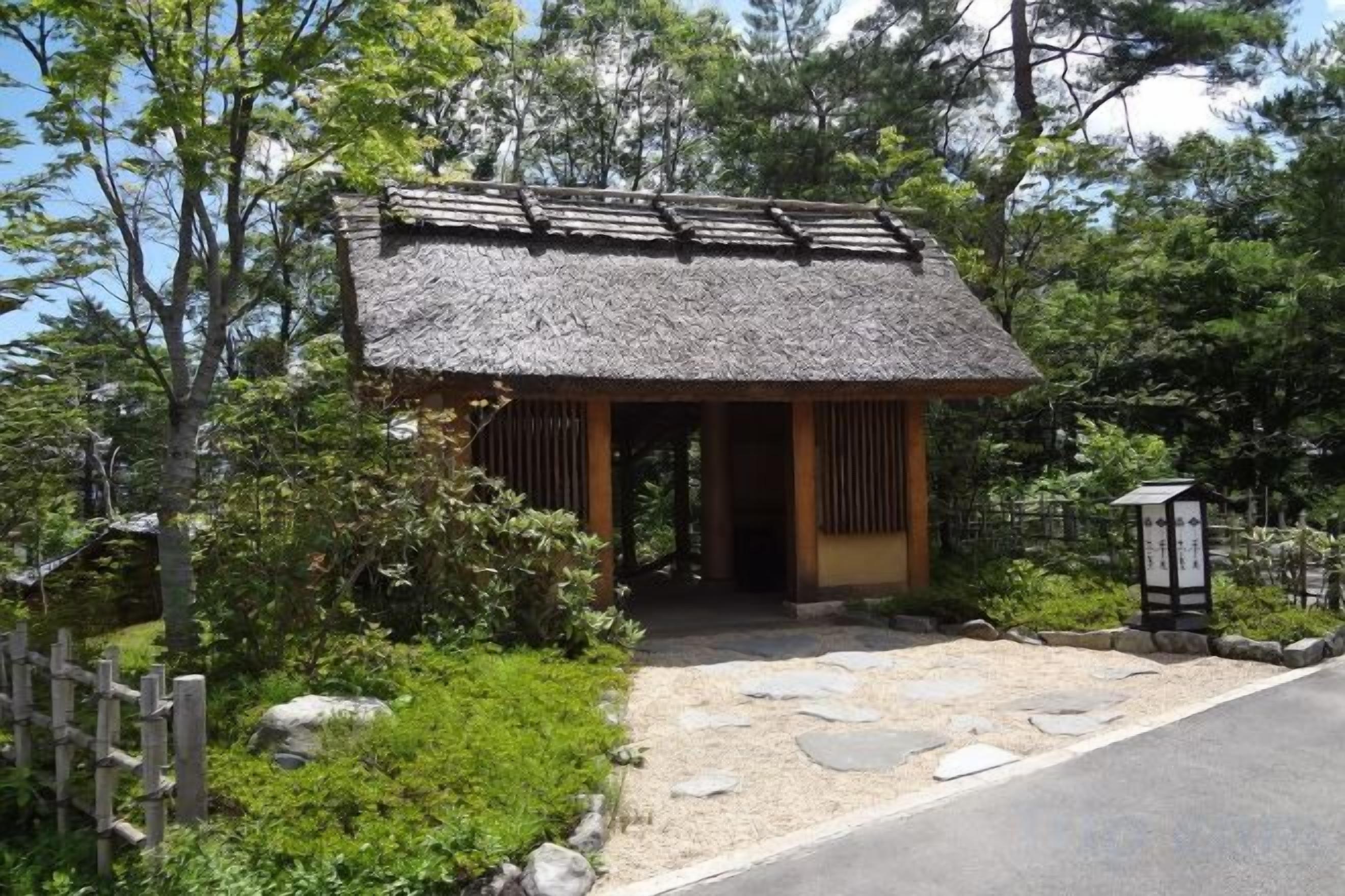 A traditional Japanese structure with a thatched roof, surrounded by greenery and a gravel path.