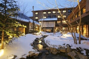 Ein mit Schnee bedeckter Innenhof mit einem Bach, mehrstöckigen Gebäuden und Außenbeleuchtung.