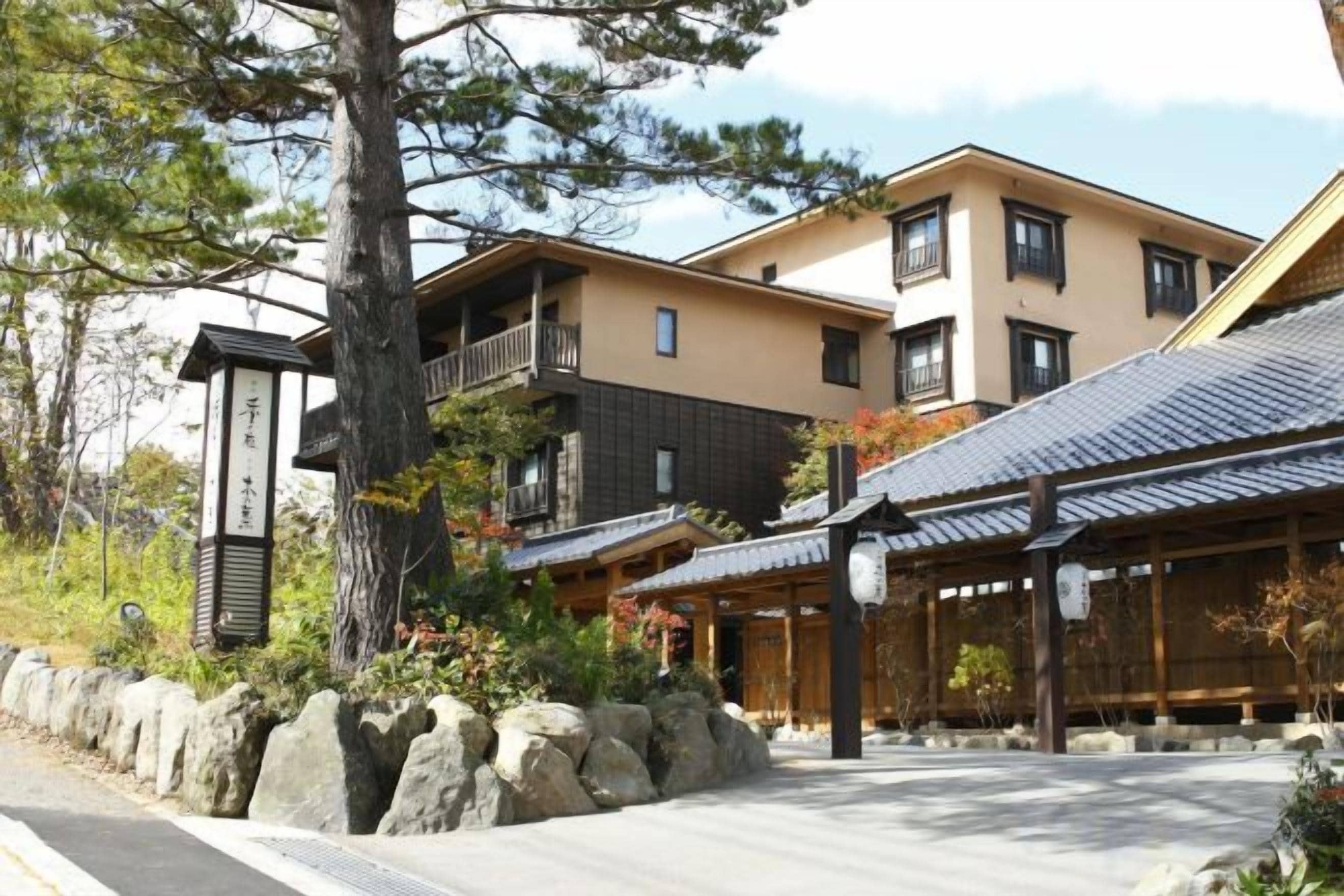 A traditional Japanese building with a stone pathway, wooden structures, and a large tree.