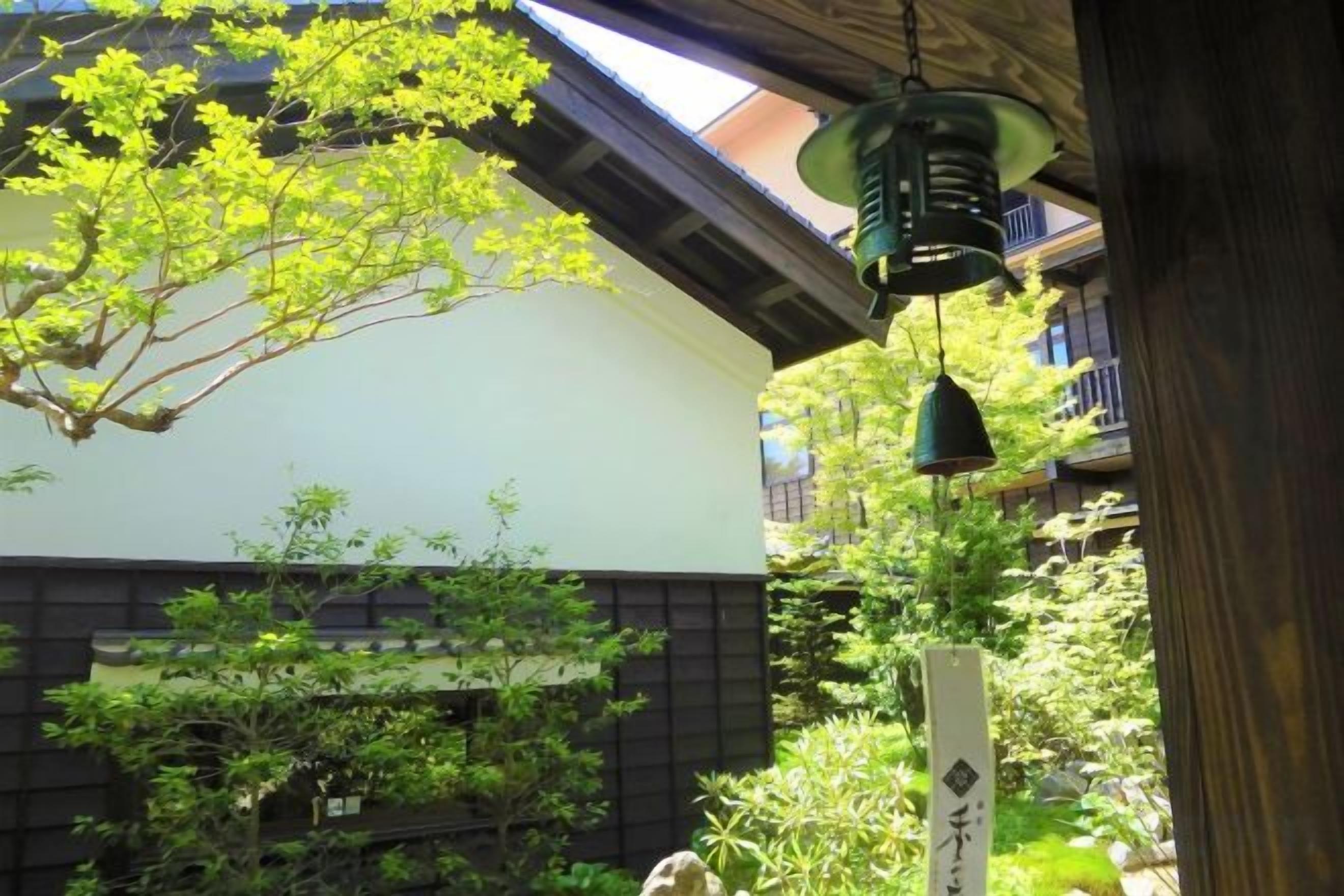 A traditional Japanese building with a sloped roof, hanging green lanterns, and a garden with various plants.