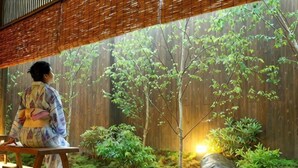 Una persona seduta su una panchina in una stanza con pavimento in legno, soffitto in bambù e vista su un giardino con alberi e rocce.