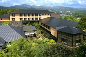 Un edificio de varios pisos con techo plano, rodeado de vegetación y montañas al fondo.