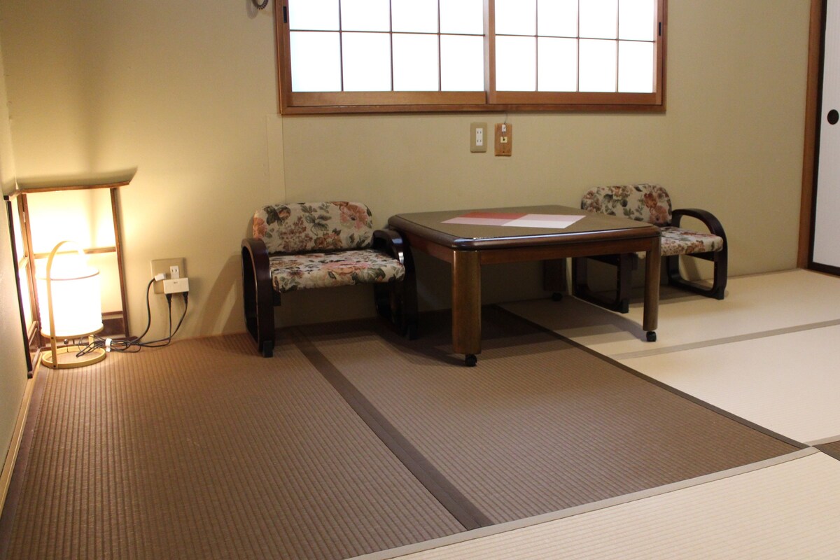 Une pièce avec un sol recouvert de tatamis, une table en bois, deux chaises à motifs floraux, une lampe et une fenêtre à carreaux.