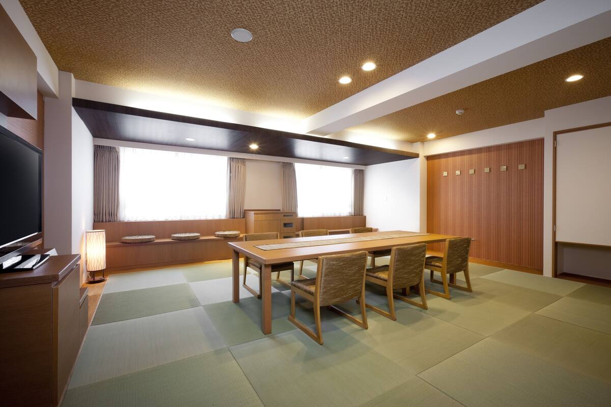 A room with a wooden dining table, chairs, a television, and a window with curtains.
