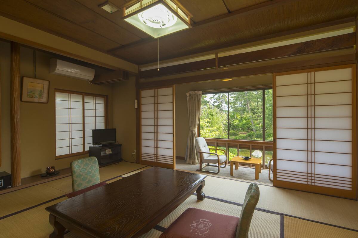 A traditional Japanese room with tatami flooring, a wooden table, a television, and sliding doors leading to a balcony with a view of greenery.