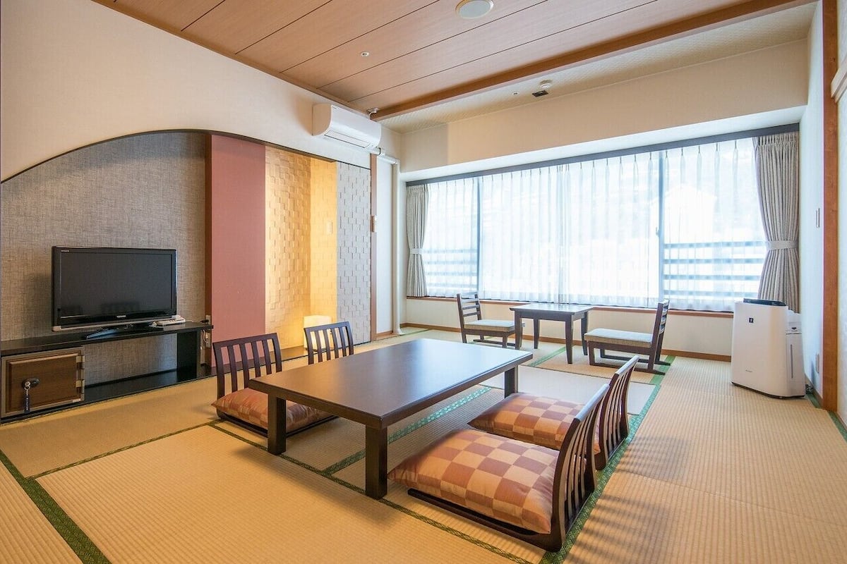 Une chambre d’hôtel avec un sol recouvert de tatamis, une télévision, une table à manger avec des chaises et une grande fenêtre avec des rideaux.