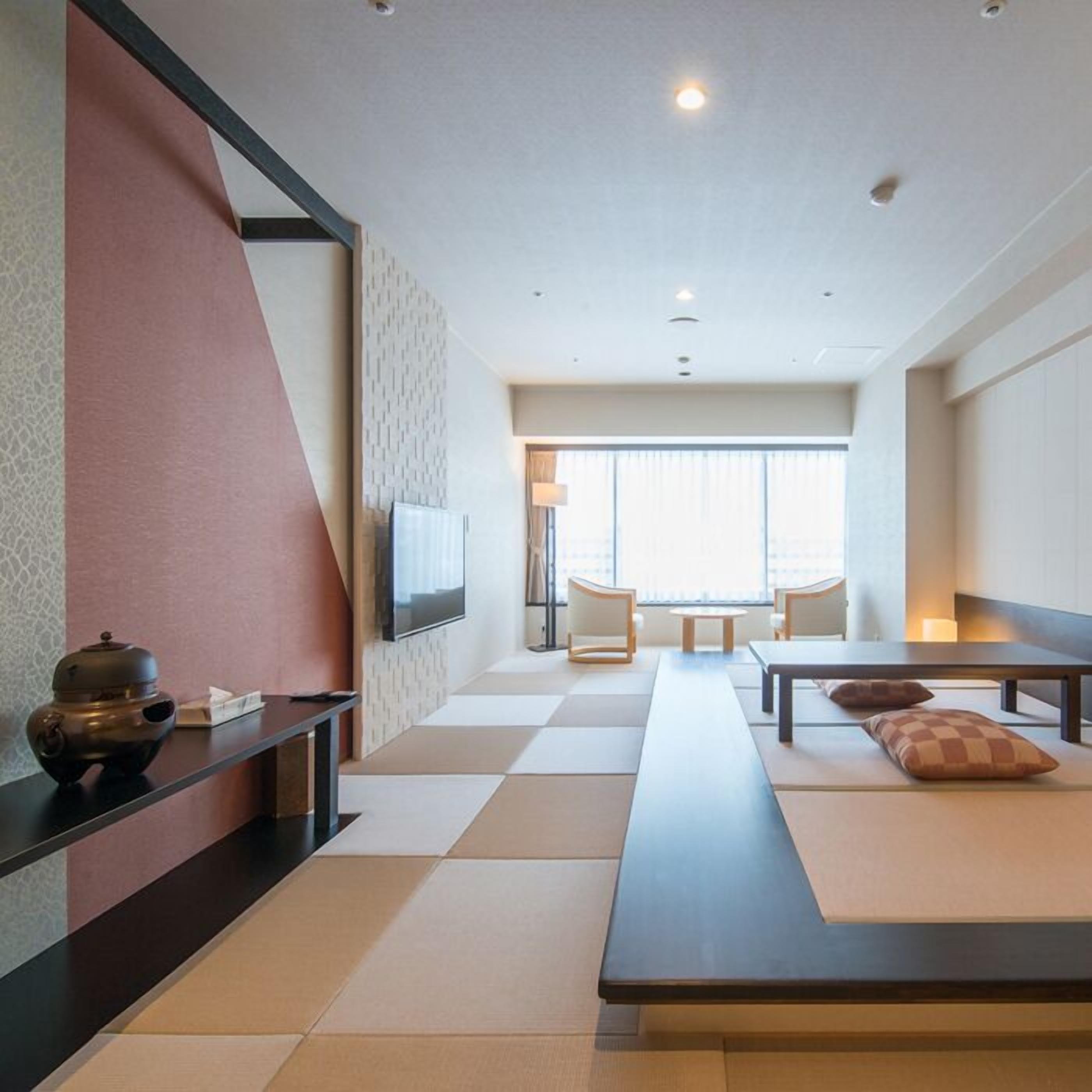 A modern hotel room with a tatami mat floor, a long table with cushions, a flat-screen TV, and a large window providing a view of the city.