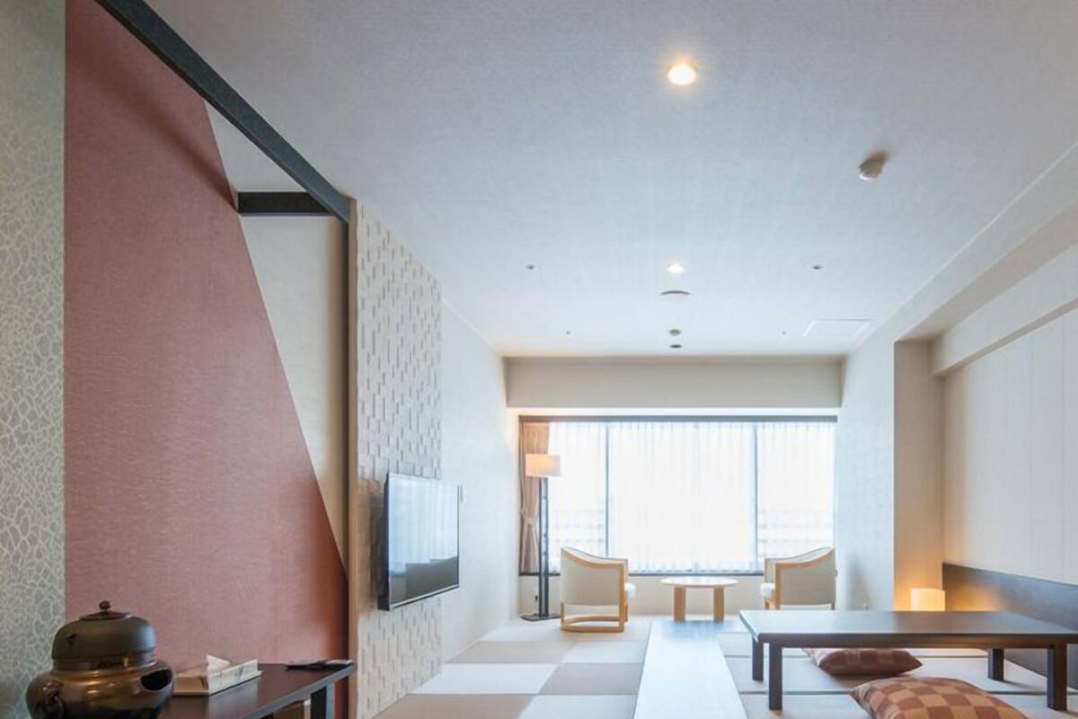 Ett modernt hotellrum med ett golv av tatamimattor, ett långt bord med kuddar, en platt-TV och ett stort fönster med utsikt över staden.