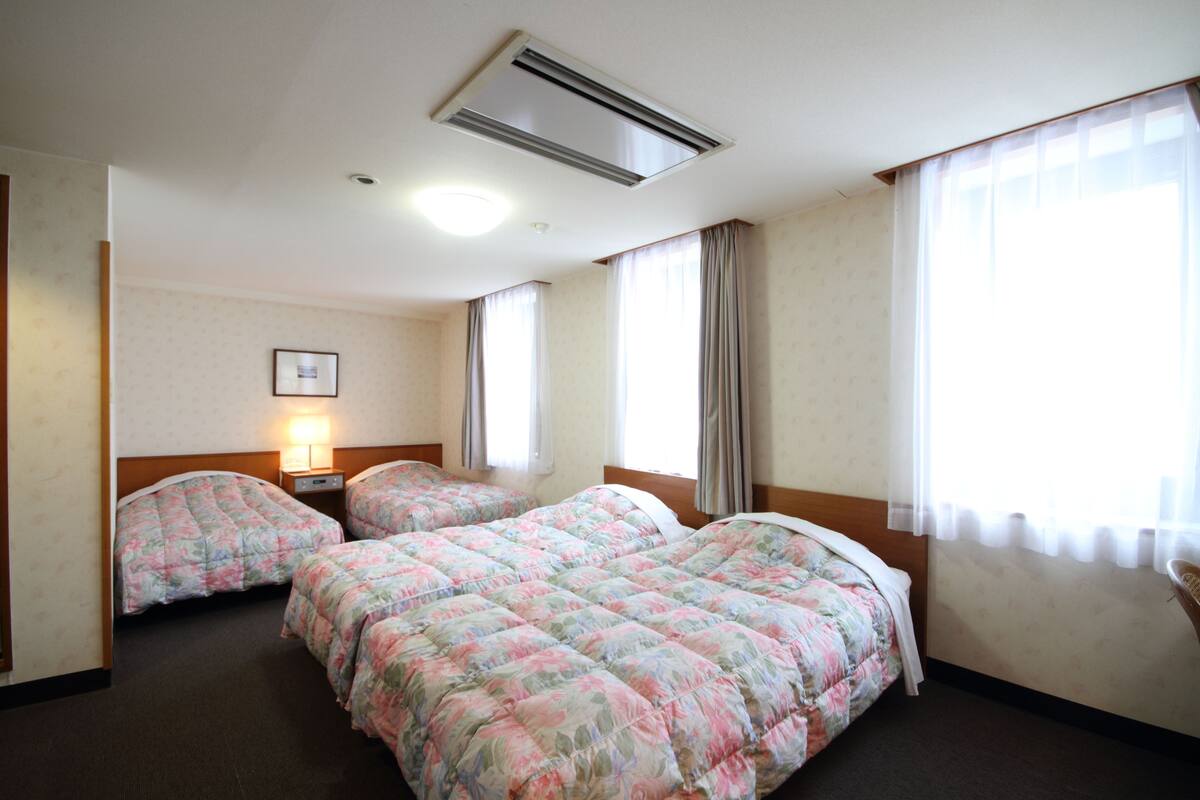 Une chambre d’hôtel avec trois lits, un ventilateur de plafond, un radiateur fixé au mur et une fenêtre avec des rideaux.