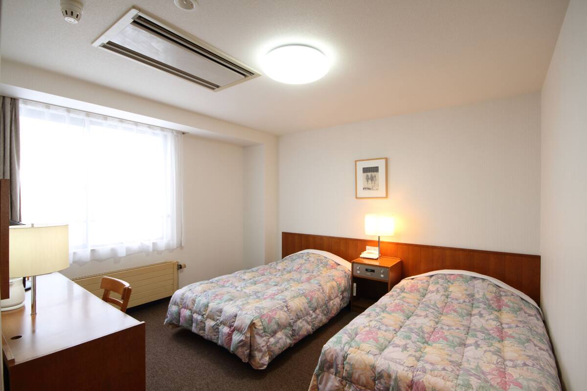 Une chambre d’hôtel avec deux lits, un bureau, une chaise, une lampe et une fenêtre avec des rideaux.