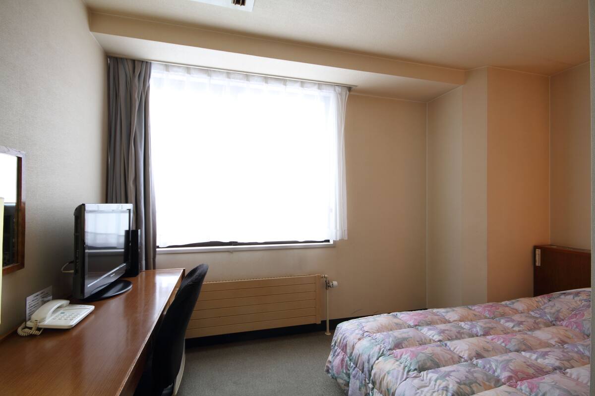 Une chambre d’hôtel comprenant un lit, un bureau avec un téléphone et une télévision, un radiateur et une fenêtre avec des rideaux.