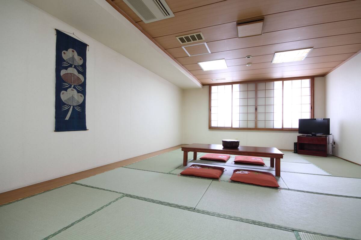 Une pièce de style japonais traditionnel, avec un sol recouvert de tatamis, une petite table en bois, des coussins, un téléviseur et un paravent en papier de riz (shoji).