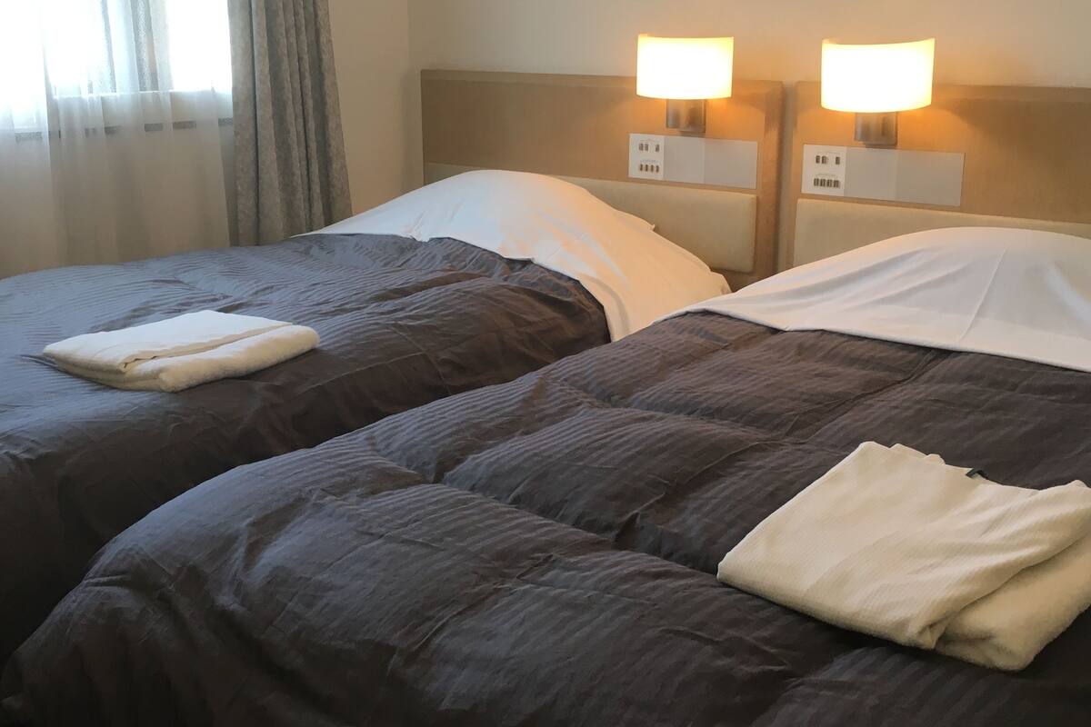 Dos camas con ropa de cama oscura y almohadas blancas, cada una con una toalla blanca doblada. Hay lámparas de pared a cada lado de las camas.