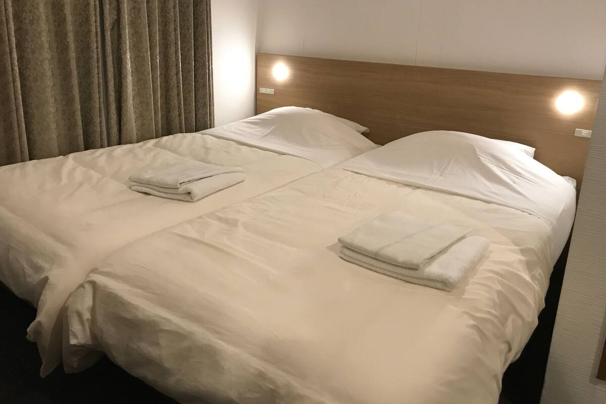 Una cama doble con sábanas blancas y dos toallas dobladas, un cabecero de madera y una lámpara fijada a la pared.