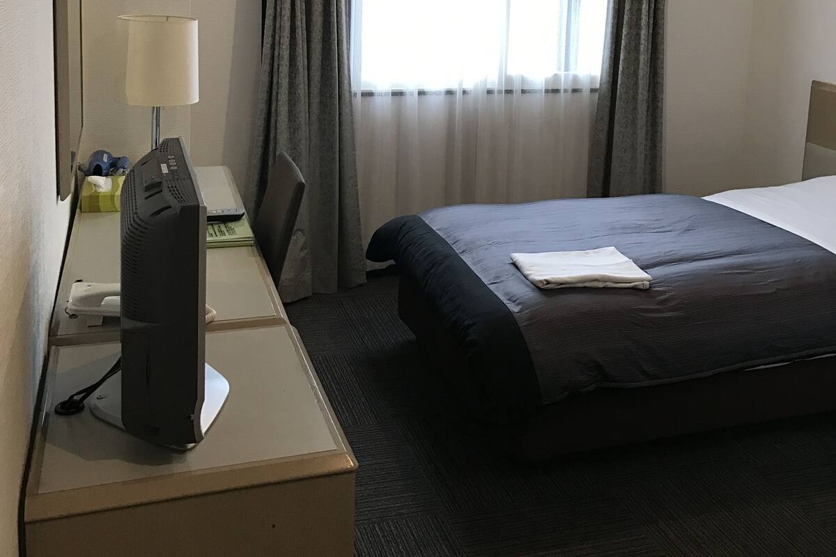 Habitación de hotel con cama, escritorio con computadora, lámpara y ventana con cortinas.