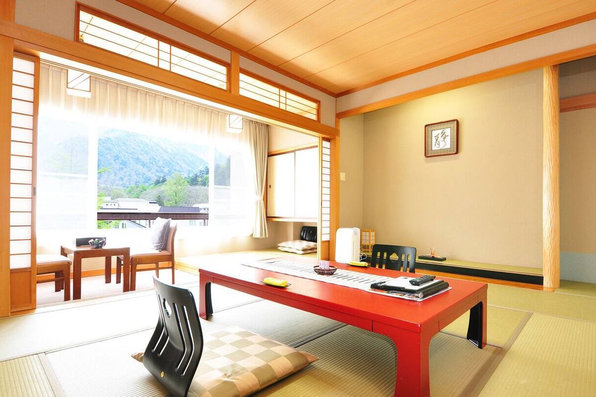 A traditional Japanese room with tatami flooring, a red table, and a view of mountains through large windows.