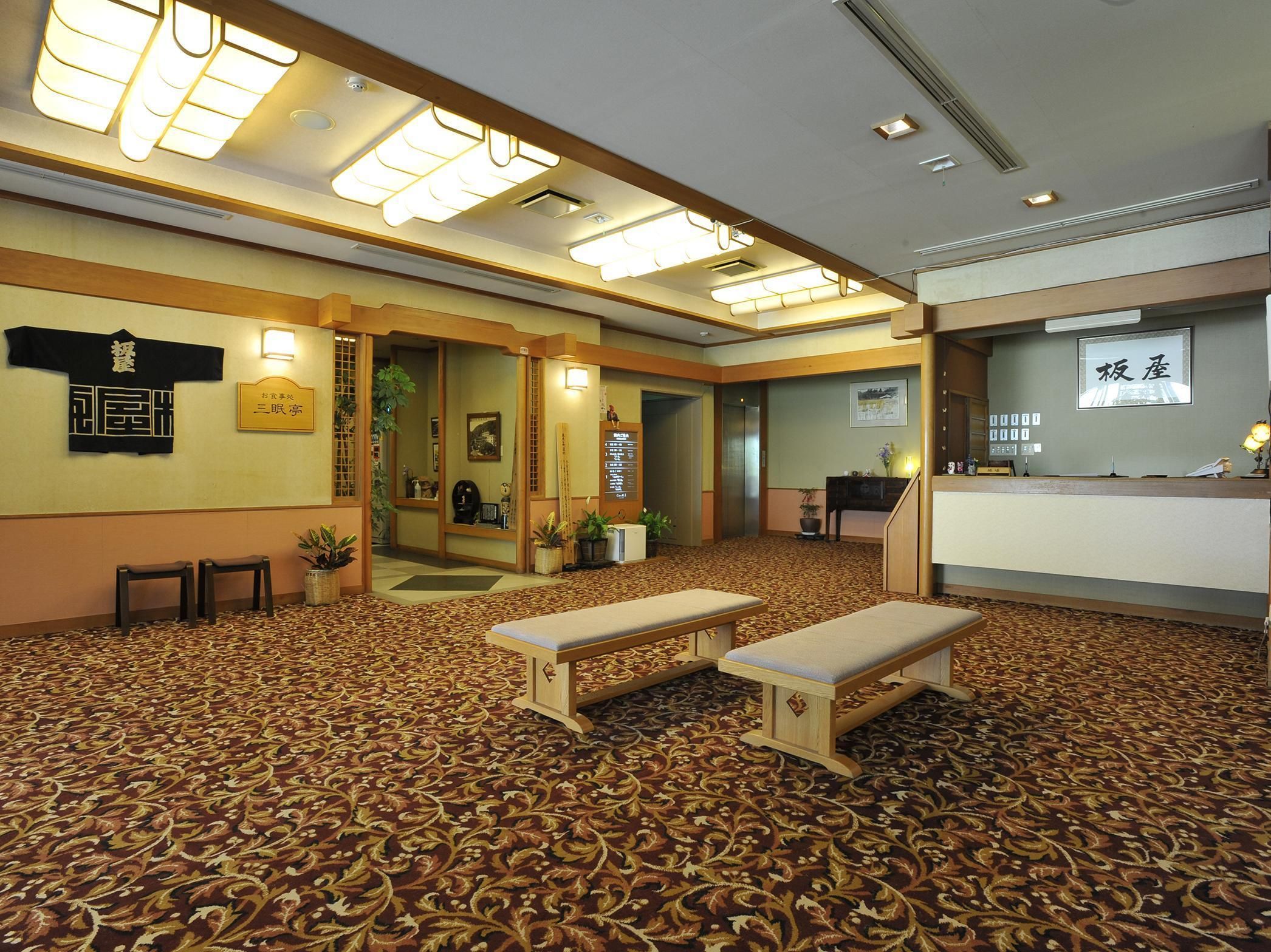 A hotel lobby with a patterned carpet, wooden benches, a reception desk, and a wall with a sign.