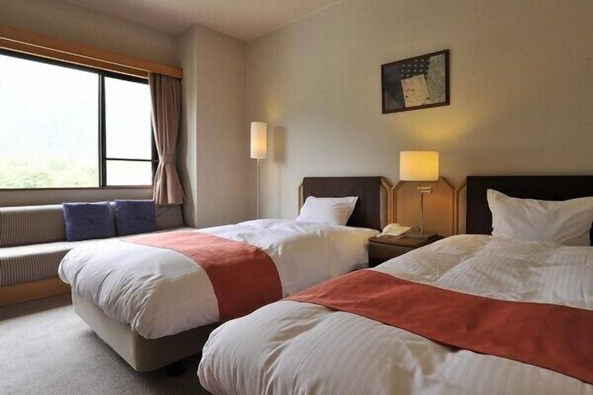 A hotel room with two beds, a window with curtains, a wall-mounted lamp, and a painting.