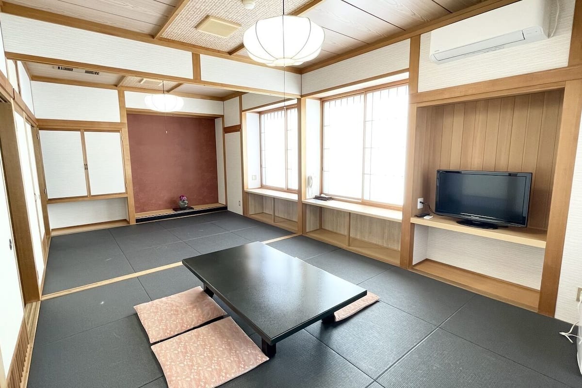 A traditional Japanese room with tatami flooring, a sliding door, a television on a shelf, and a ceiling fan.
