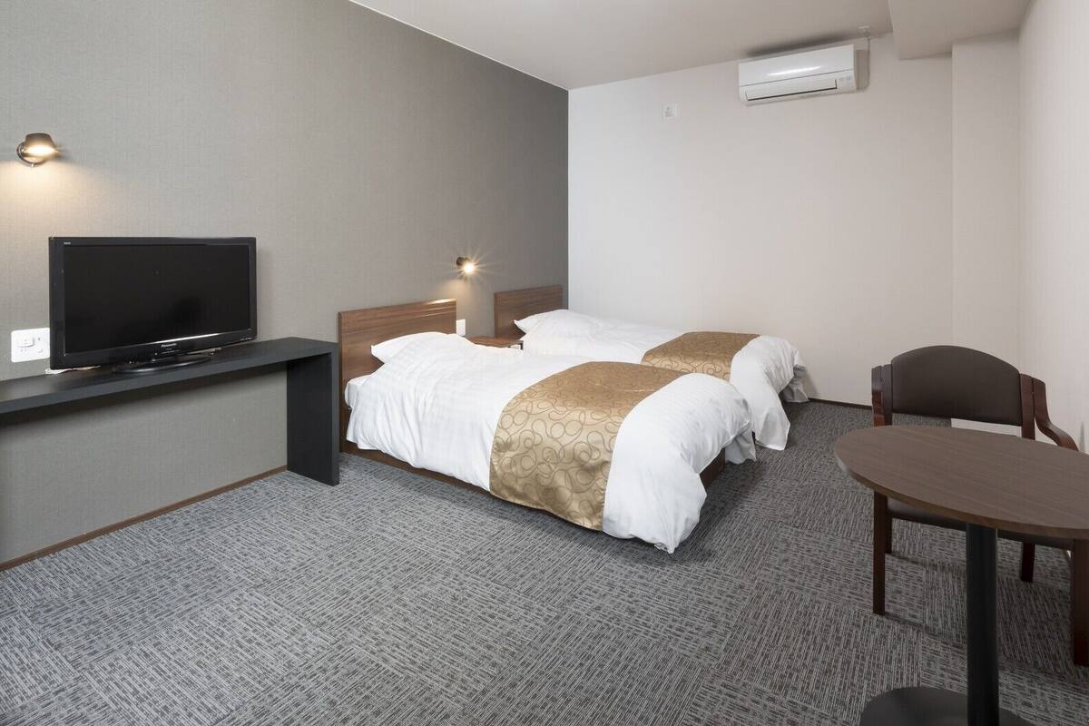 A hotel room with a TV, two beds, a desk, a chair, and an air conditioner.