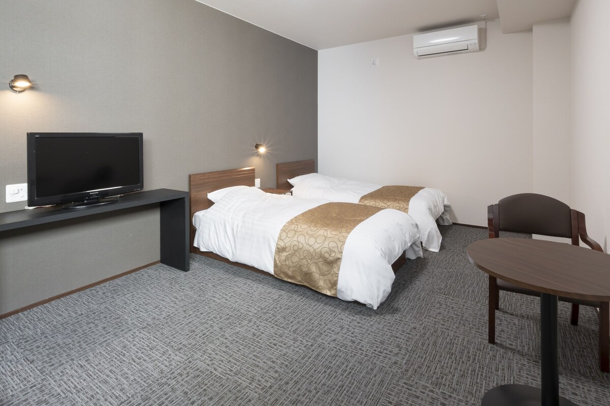 Habitación de hotel con televisión, dos camas, escritorio, silla y aire acondicionado.