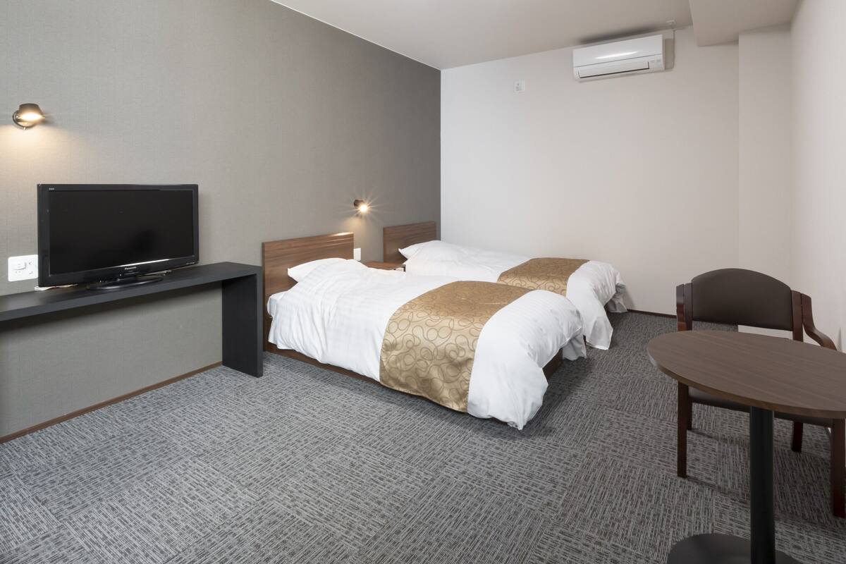 A hotel room with a TV, two beds, a desk, a chair, and an air conditioning unit.