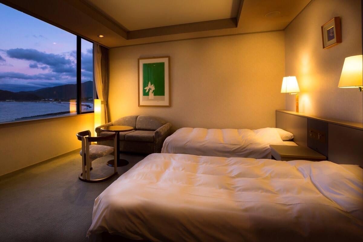 A hotel room with two beds, a small table, a chair, and a view of the sea.